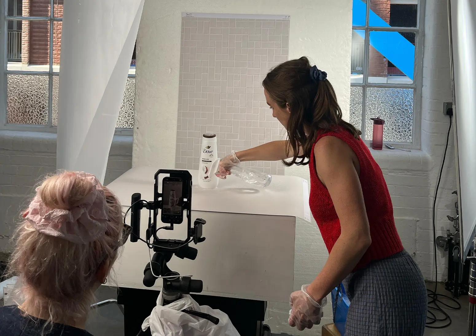 A small crew filming a product shot as a person pours liquid beside a Dove bottle on a white tabletop, with a smartphone mounted on a tripod capturing the setup.